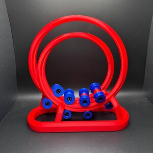 May include: A red, 3D-printed abacus-style toy with two large circular frames and a series of blue and red beads. The base is also red, and the toy is set against a dark background.