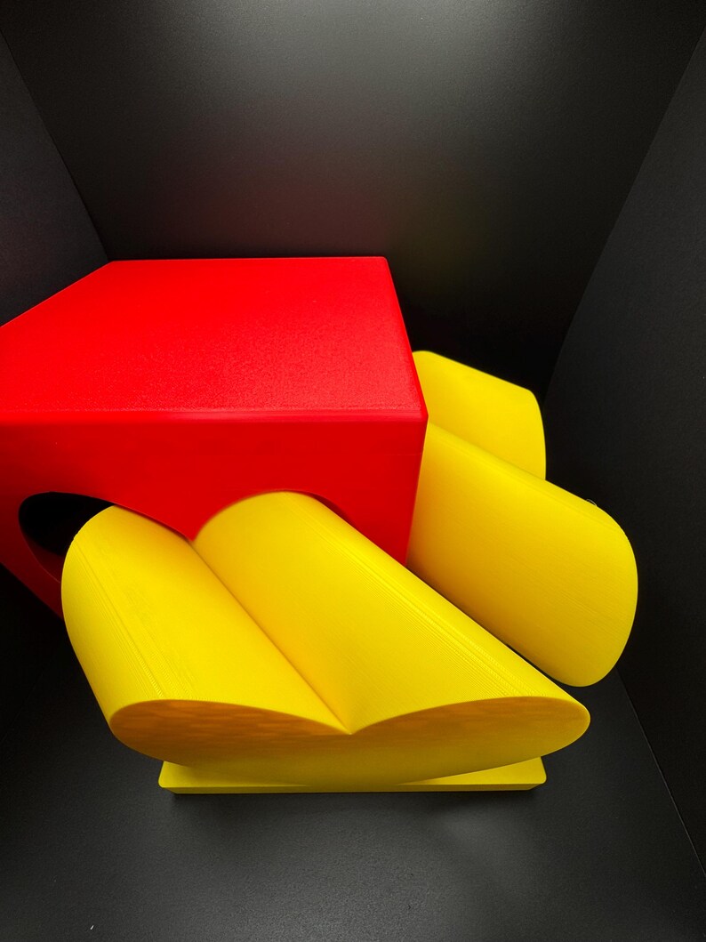 Pode incluir: Um cubo vermelho com um objeto amarelo em forma de cora&ccedil;&atilde;o no topo. O objeto em forma de cora&ccedil;&atilde;o &eacute; feito de duas pe&ccedil;as curvas que se juntam.