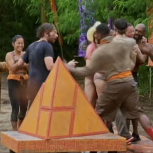 May include: A group of people stand around a large, orange and red pyramid-shaped structure. The pyramid is on a wooden platform with intricate carvings. The people are wearing casual clothing and appear to be participating in a game or challenge.