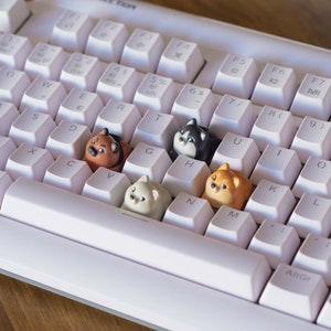 May include: A white mechanical keyboard with custom keycaps shaped like cartoon dogs. The keycaps are various colors, including brown, black, and cream. The keyboard has a standard layout with white keys and black lettering.