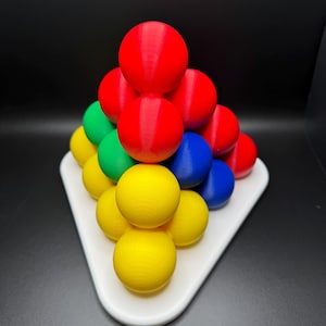 May include: A pyramid-shaped puzzle made of colorful balls. The balls are red, yellow, green, and blue. The puzzle is on a white triangular base.