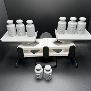 May include: A white balance scale with gray weights numbered 1, 2, 3, 4, 5, 6, 7, and 8. The scale is on a black surface.
