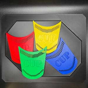 May include: Four colorful plastic cup-shaped objects in red, yellow, green, and blue, each with the word "CUP" printed on them. The objects are arranged on a black octagonal base.