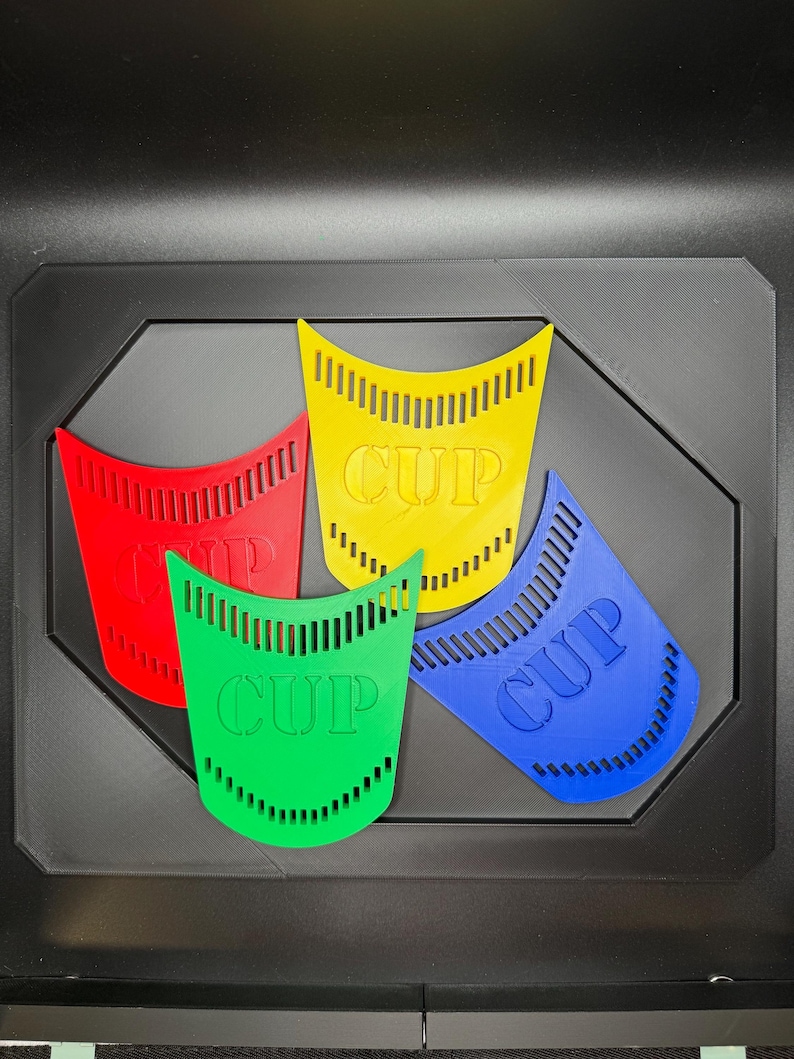 May include: Four colorful plastic cup-shaped objects in red, yellow, green, and blue, each with the word "CUP" printed on them. The objects are arranged on a black octagonal base.