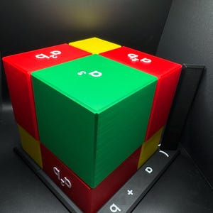 May include: A colorful geometric puzzle with red, green, and yellow blocks. White text with mathematical symbols is visible on the blocks and base. The puzzle is set against a dark background.