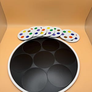 May include: A black and white game board with seven black circular spaces. Seven white circular game pieces with colorful dots are arranged above the board. The dots are in various colors, including blue, green, and yellow.