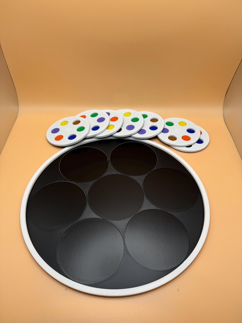 May include: A black and white game board with seven black circular spaces. Seven white circular game pieces with colorful dots are arranged above the board. The dots are in various colors, including blue, green, and yellow.