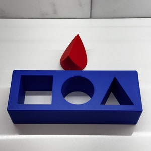 May include: A blue plastic block with cutouts for a square, circle, and triangle. A red plastic teardrop shape sits on top of the block.