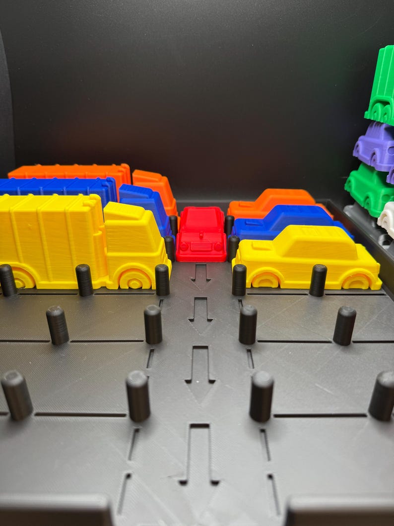 May include: A selection of brightly coloured toy cars, including yellow, orange, blue, and red, arranged on a grey play surface. The cars are 3D-printed and appear to be part of a children's toy set. The background is black.