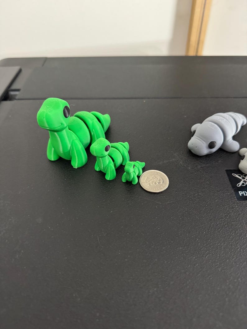Puede incluir: Tres figuras de dinosaurio impresas en 3D de diferentes tama&ntilde;os, la m&aacute;s grande es aproximadamente del tama&ntilde;o de una moneda de 20 c&eacute;ntimos de euro. Los dinosaurios tienen ojos grandes y est&aacute;n segmentados.