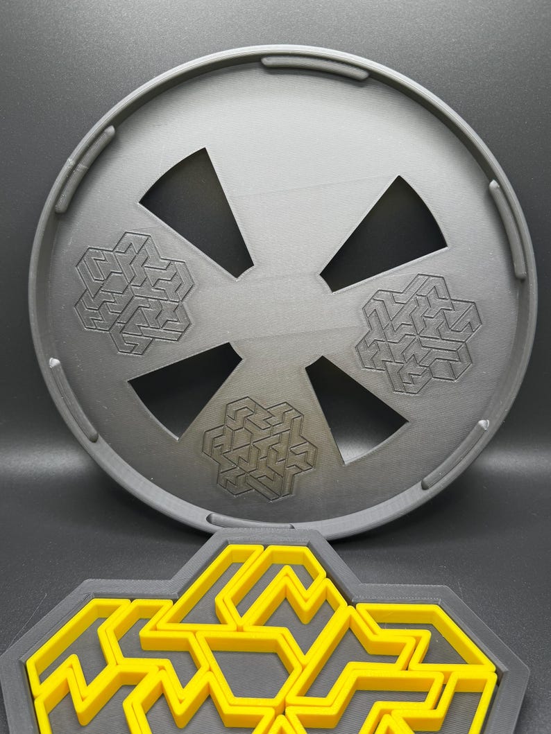 May include: A gray plastic circular puzzle with three interlocking geometric shapes. The puzzle is designed to be disassembled and reassembled. The puzzle is shown on a black surface.