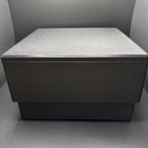 May include: A black square box with a textured lid. The box is closed.