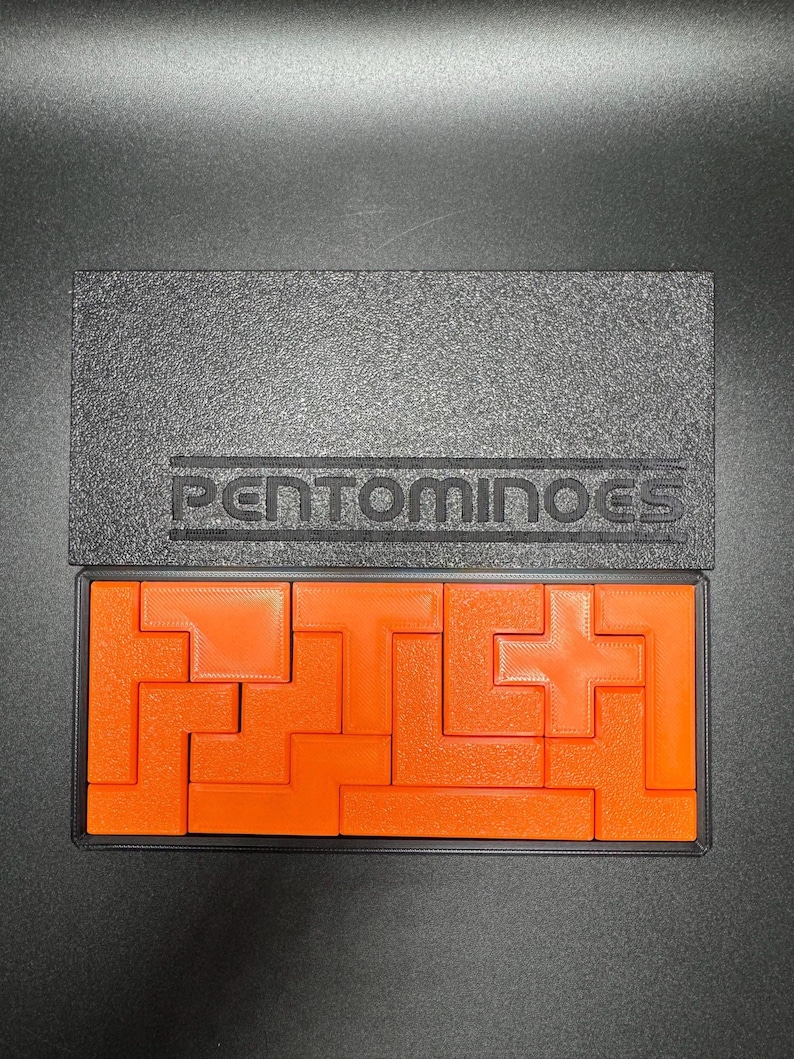 May include: A set of orange plastic pentominoes in a black box with the word "Pentominoes" printed on the lid. The pieces are arranged in a puzzle pattern.