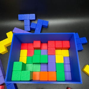 May include: A blue rectangular container filled with colorful, geometric puzzle pieces. The pieces are various shapes and colors, including red, yellow, green, orange, and purple. Some pieces are outside the container.
