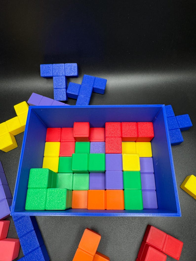 May include: A blue rectangular container filled with colorful, geometric puzzle pieces. The pieces are various shapes and colors, including red, yellow, green, orange, and purple. Some pieces are outside the container.