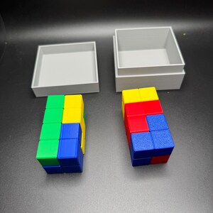 Puede incluir: Dos cajas grises con tapas, una abierta y otra cerrada. La caja abierta contiene una torre de bloques de colores, rojo, azul, amarillo y verde. La caja cerrada contiene una torre de bloques similar.