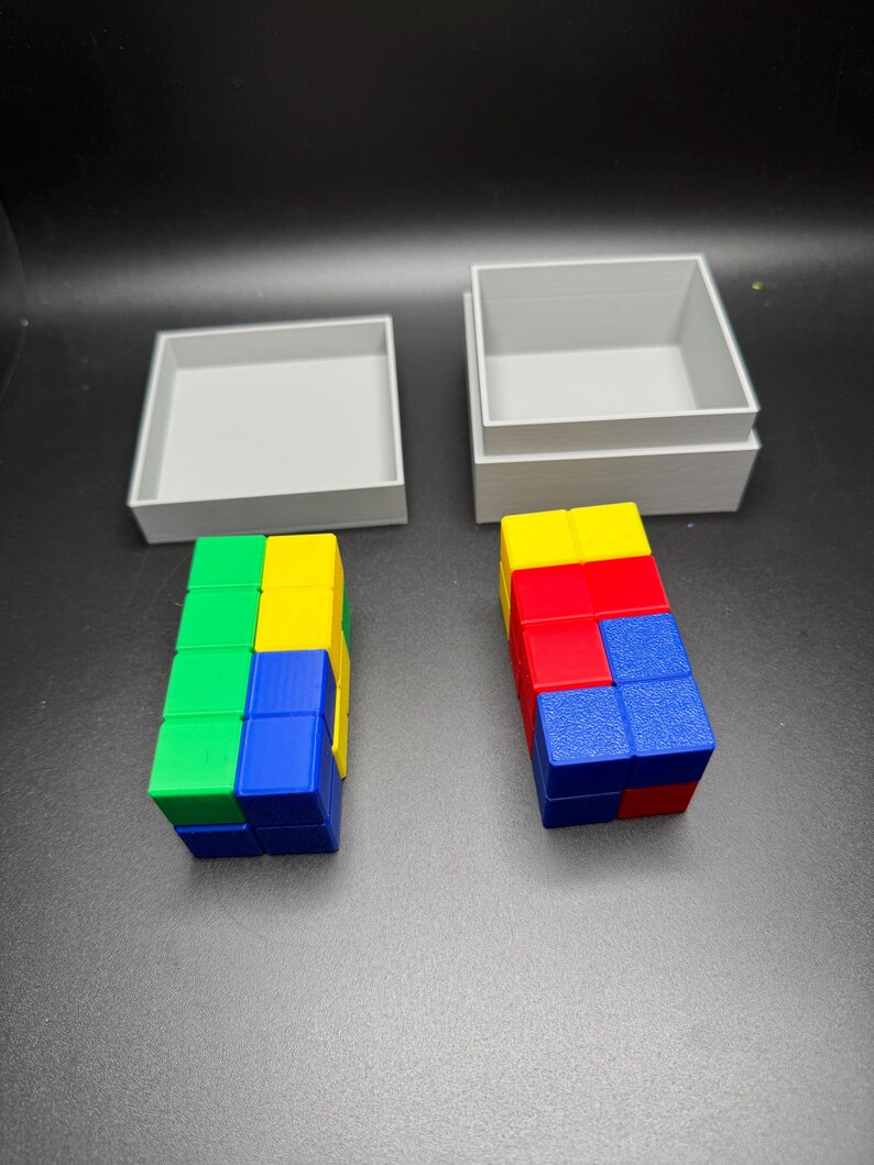 Puede incluir: Dos cajas grises con tapas, una abierta y otra cerrada. La caja abierta contiene una torre de bloques de colores, rojo, azul, amarillo y verde. La caja cerrada contiene una torre de bloques similar.