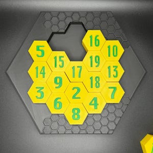Puede incluir: Un rompecabezas hexagonal con fichas hexagonales amarillas numeradas del 2 al 19. Las fichas est&aacute;n dispuestas en un patr&oacute;n de panal con una ficha que falta en el centro.