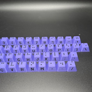 May include: A set of purple keyboard keys with black lettering and braille dots. The keys include numbers, letters, punctuation marks, and symbols.