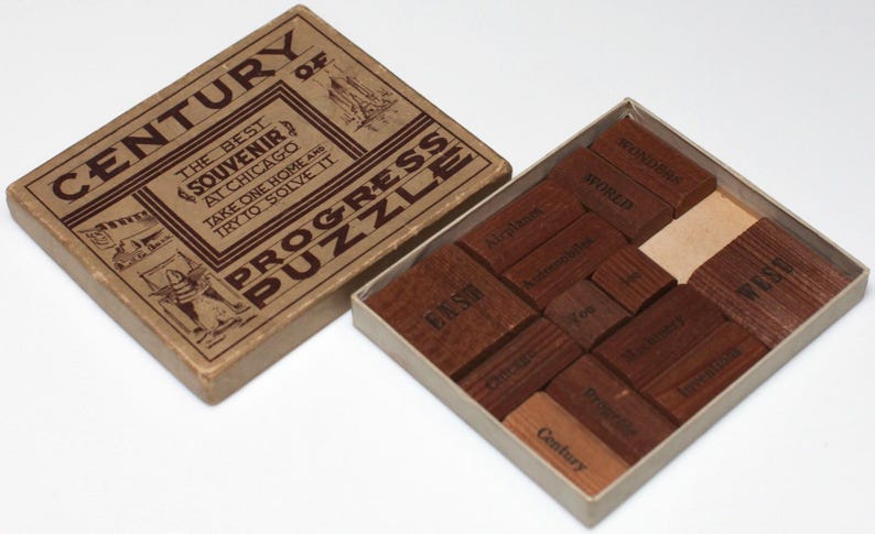 May include: A wooden box with a brown paper label that reads "Century of Progress Puzzle" and "The Best Souvenir at Chicago. Take one home and try to solve it." The box contains a wooden puzzle with 12 pieces, each with a different word printed on it.