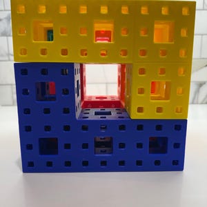 May include: A three-dimensional structure made of interlocking plastic cubes. The top layer is yellow, the middle layer is blue and red, and the bottom layer is blue. The structure has a hollow center.