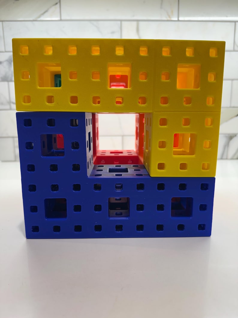 May include: A three-dimensional structure made of interlocking plastic cubes. The top layer is yellow, the middle layer is blue and red, and the bottom layer is blue. The structure has a hollow center.