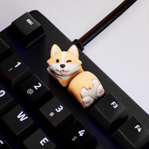 Op de afbeelding: Een zwart toetsenbord met een aangepaste corgi-vormige keycap. De keycap is bruin, wit en bruin, met de corgi die knipoogt en zijn tong uitsteekt. Het toetsenbord heeft zwarte toetsen met witte letters.