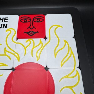 May include: A close-up of a puzzle with white and red square tiles. The tiles feature yellow flame designs and a red sun face. The text "THE SUN" is visible on a white tile. The puzzle is set in a black tray.