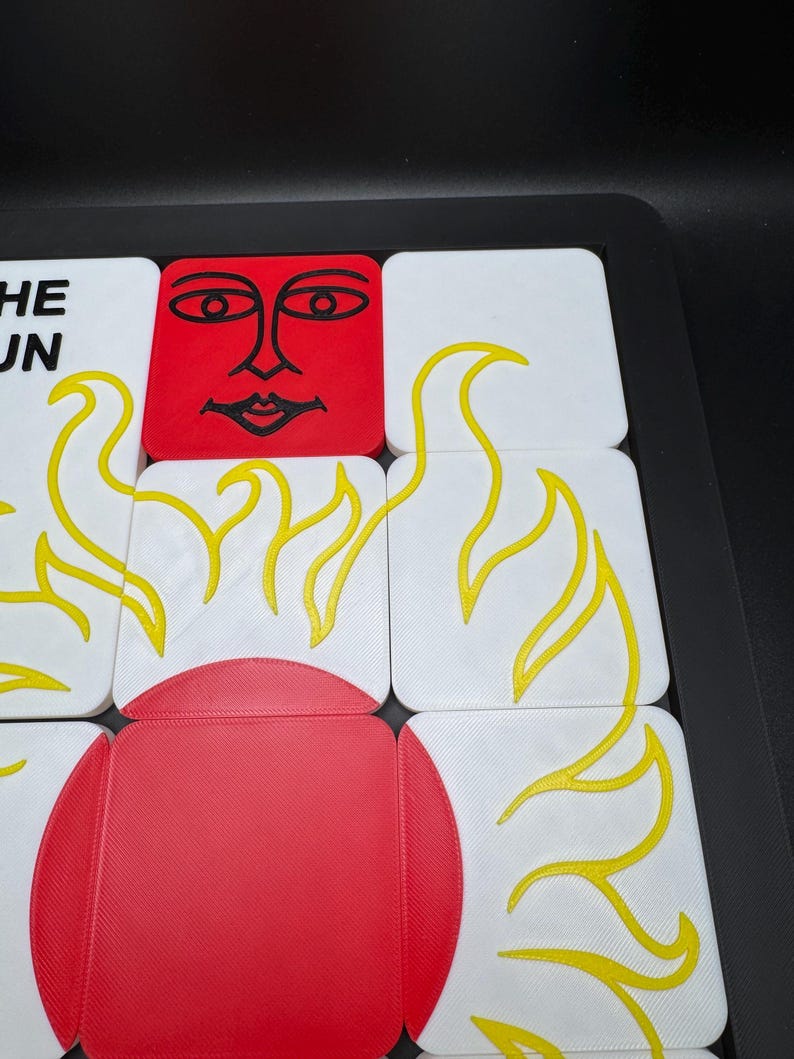 May include: A close-up of a puzzle with white and red square tiles. The tiles feature yellow flame designs and a red sun face. The text "THE SUN" is visible on a white tile. The puzzle is set in a black tray.