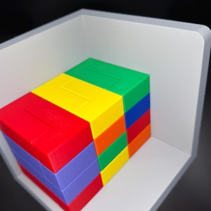 May include: A multi-colored plastic puzzle block set featuring stacked rectangular pieces in red, yellow, green, blue, orange, and purple. The blocks are arranged in a compact, rectangular formation and are positioned inside a light gray corner shelf.