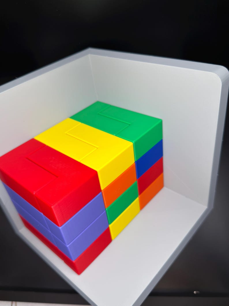 May include: A multi-colored plastic puzzle block set featuring stacked rectangular pieces in red, yellow, green, blue, orange, and purple. The blocks are arranged in a compact, rectangular formation and are positioned inside a light gray corner shelf.