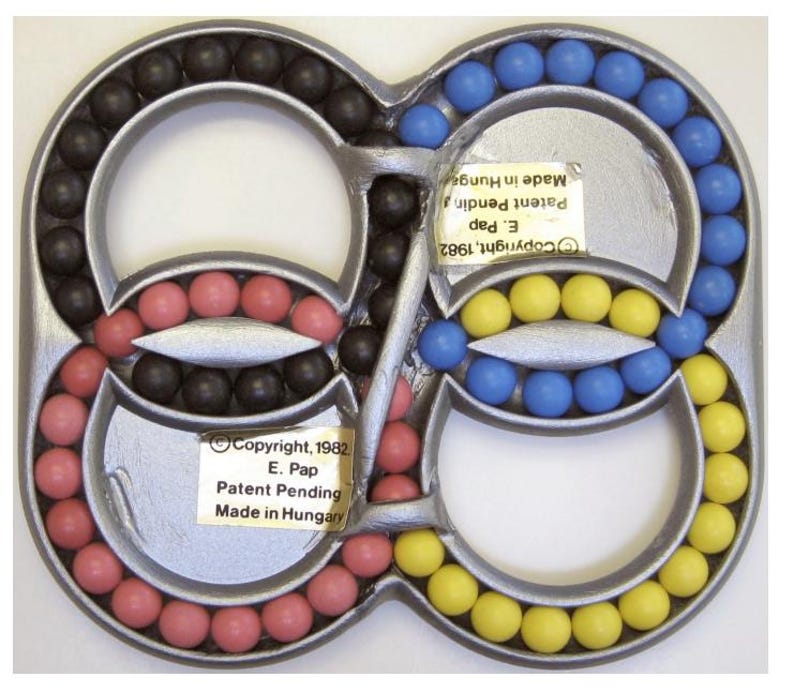 May include: A silver-colored puzzle toy with an infinity symbol design. The toy features colored balls in black, pink, blue, and yellow. The text on the toy reads "Copyright 1982 E. Pap Patent Pending Made in Hungary."