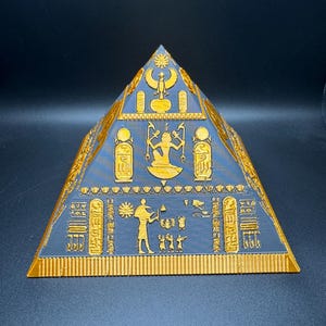 Puede incluir: Una pir&aacute;mide impresa en 3D con intrincados jerogl&iacute;ficos y figuras doradas sobre una superficie azul oscuro. El dise&ntilde;o de la pir&aacute;mide recuerda a la arquitectura del antiguo Egipto, con tallas detalladas y una forma sim&eacute;trica. Es un objeto decorativo.