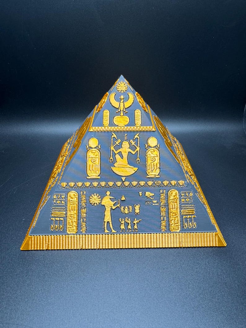 Puede incluir: Una pir&aacute;mide impresa en 3D con intrincados jerogl&iacute;ficos y figuras doradas sobre una superficie azul oscuro. El dise&ntilde;o de la pir&aacute;mide recuerda a la arquitectura del antiguo Egipto, con tallas detalladas y una forma sim&eacute;trica. Es un objeto decorativo.