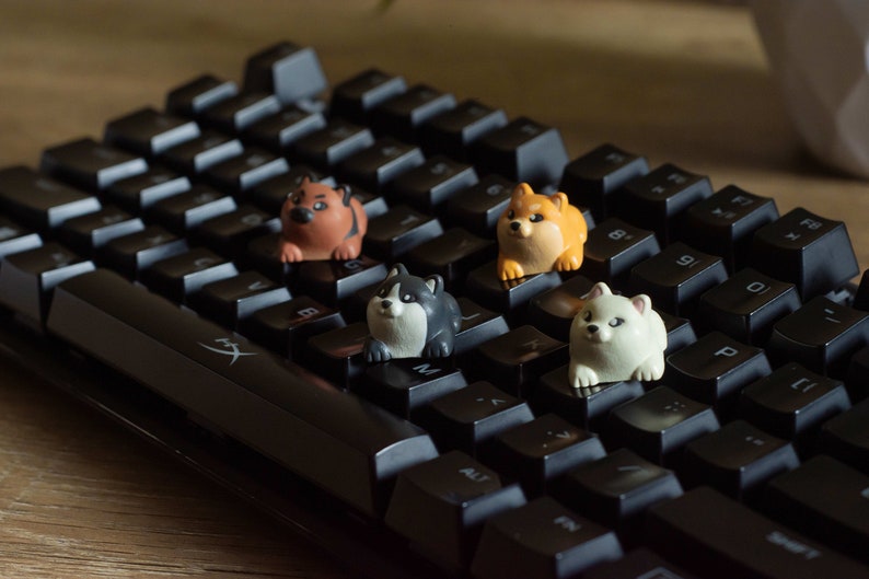 May include: A black mechanical keyboard with four colorful Shiba Inu keycaps. The keycaps are orange, white, black and brown, and are shaped like cartoon dogs. The keyboard is on a wooden surface.