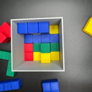 Puede incluir: Una caja gris llena de bloques de madera de colores. Los bloques est&aacute;n dispuestos en un patr&oacute;n y son de madera azul, verde, amarilla y roja. La caja est&aacute; sobre una superficie negra.