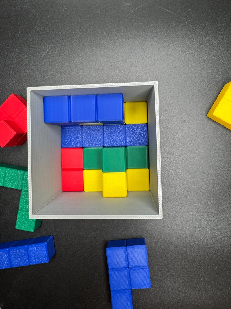 Puede incluir: Una caja gris llena de bloques de madera de colores. Los bloques est&aacute;n dispuestos en un patr&oacute;n y son de madera azul, verde, amarilla y roja. La caja est&aacute; sobre una superficie negra.