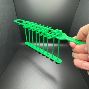 Pode incluir: Brinquedo de pl&aacute;stico verde com uma s&eacute;rie de la&ccedil;os e bolas pretas. O brinquedo &eacute; segurado na m&atilde;o.