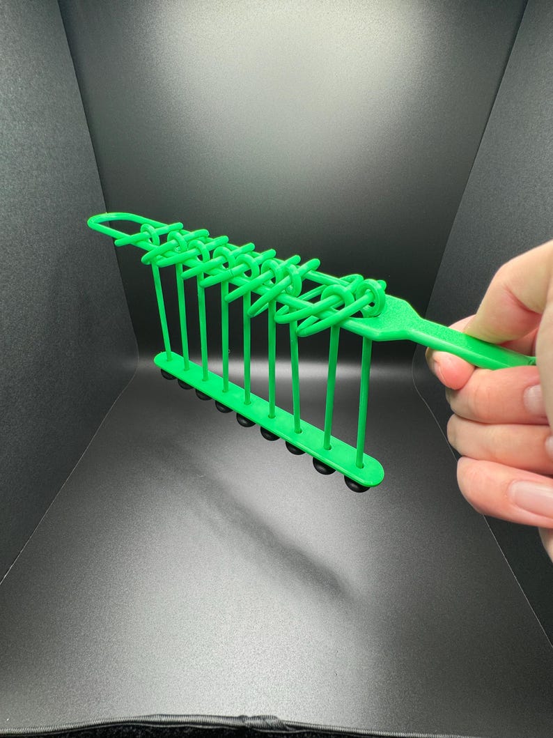 Pode incluir: Brinquedo de pl&aacute;stico verde com uma s&eacute;rie de la&ccedil;os e bolas pretas. O brinquedo &eacute; segurado na m&atilde;o.