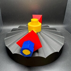May include: A 3D-printed abstract sculpture featuring a black, fan-shaped base. A yellow cylindrical structure is topped with a red triangular prism. A blue hexagonal shape with a yellow center and a red square are also present.