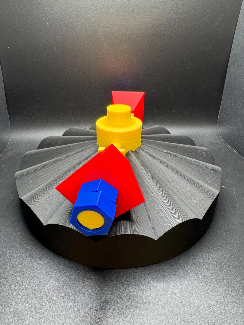 May include: A 3D-printed abstract sculpture featuring a black, fan-shaped base. A yellow cylindrical structure is topped with a red triangular prism. A blue hexagonal shape with a yellow center and a red square are also present.