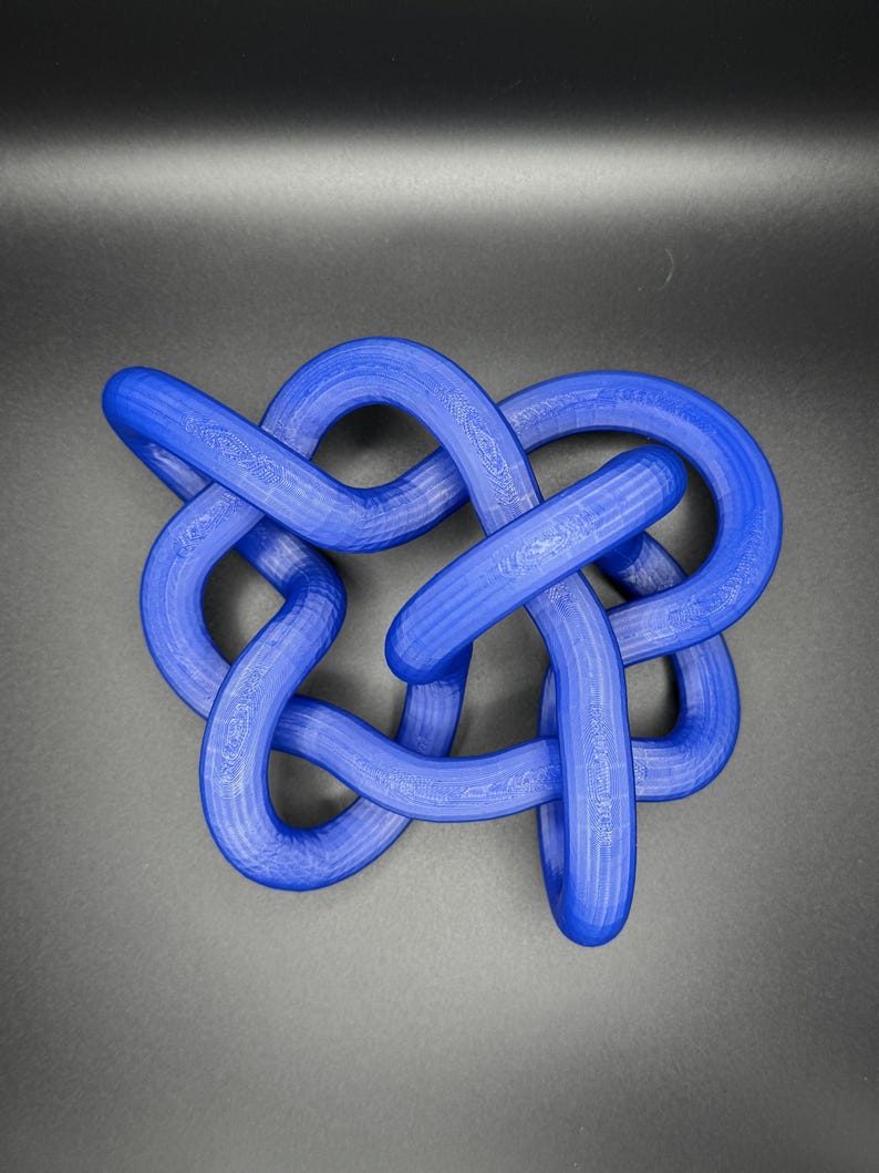 K&ouml;nnte beinhalten: Eine blaue, 3D-gedruckte Knotenskulptur. Der Knoten besteht aus einem einzigen St&uuml;ck Plastik und ist in einem komplexen Muster gebunden.