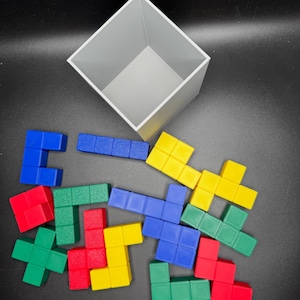 Puede incluir: Un conjunto de bloques de Tetris de pl&aacute;stico de colores rojo, azul, verde y amarillo. Los bloques est&aacute;n esparcidos sobre una superficie negra, con un contenedor en forma de cubo gris en el fondo.