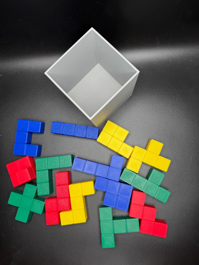 Puede incluir: Un conjunto de bloques de Tetris de pl&aacute;stico de colores rojo, azul, verde y amarillo. Los bloques est&aacute;n esparcidos sobre una superficie negra, con un contenedor en forma de cubo gris en el fondo.