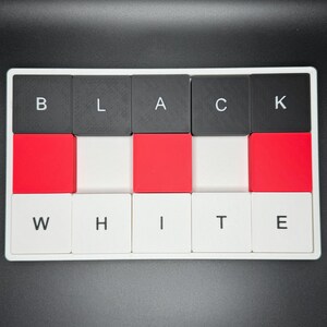 May include: A rectangular puzzle with the words "BLACK" and "WHITE" spelled out in individual letter tiles. The tiles alternate between black, red, and white, creating a visually striking contrast. The letters are in a sans-serif font.
