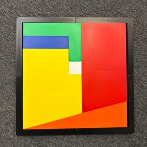 May include: A square puzzle board featuring a black frame containing various geometric shapes in bright colours. The arrangement includes a large yellow rectangle, a red rectangle, a green L-shaped piece, a blue rectangle, an orange triangle, and a small white square.
