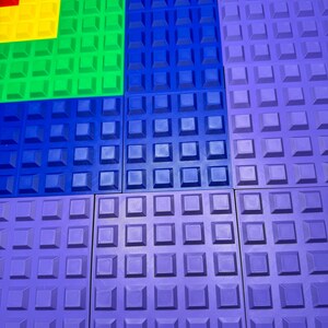 May include: A collection of colorful, textured square tiles in shades of red, yellow, green, blue, and purple. Each tile features a grid of raised square studs, creating a tactile surface. The tiles are arranged in a geometric pattern.