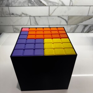 May include: A black cube with a colorful pattern on the top. The pattern is made up of squares in shades of purple, orange, and yellow.