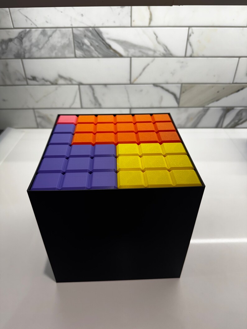 May include: A black cube with a colorful pattern on the top. The pattern is made up of squares in shades of purple, orange, and yellow.