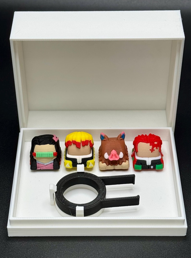 May include: A white box with a black ring and four colorful 3D printed keycaps featuring characters from the anime Demon Slayer. The keycaps are in a row, from left to right: Nezuko Kamado, Tanjiro Kamado, Inosuke Hashibira, and Zenitsu Agatsuma.